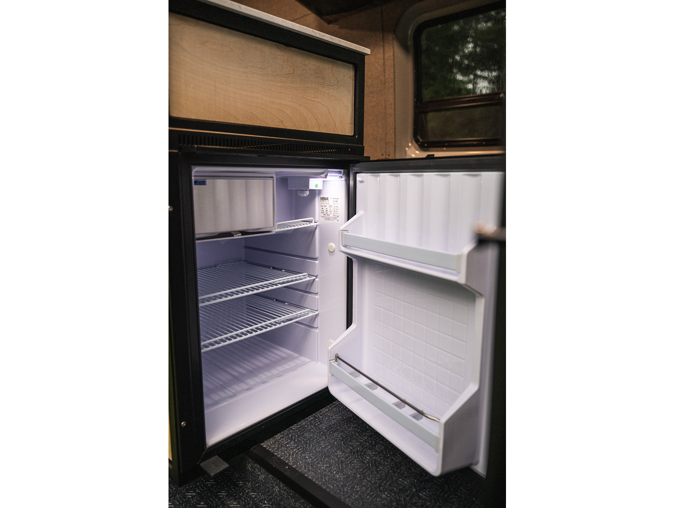 RV rental compact fridge, door open. Interior shows wire shelves, door storage, and small top freezer compartment. Ready for your adventure.