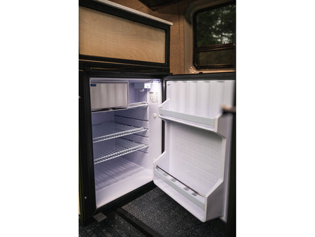 RV rental compact fridge, door open. Interior shows wire shelves, door storage, and small top freezer compartment. Ready for your adventure.