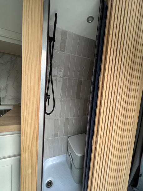 An RV rental features a compact wet bath with black shower, grey tiled walls, and a portable composting toilet.