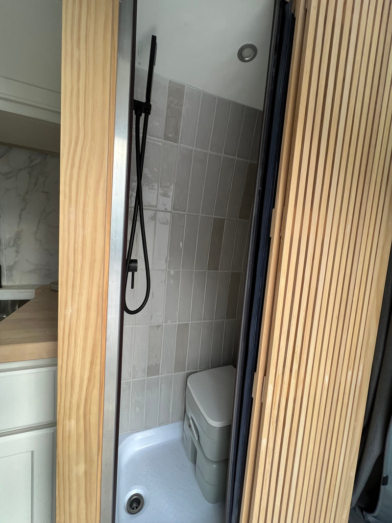 An RV rental features a compact wet bath with black shower, grey tiled walls, and a portable composting toilet. — photo 8