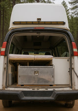 Rear view of an RV rental camper van with its back hatch open, revealing a built-in bed, storage drawer, and cooler inside.