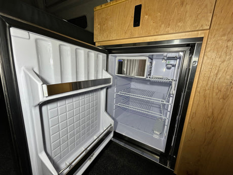 RV rental: Open, empty compact refrigerator with freezer and shelves, integrated into light wood cabinetry for efficient storage.