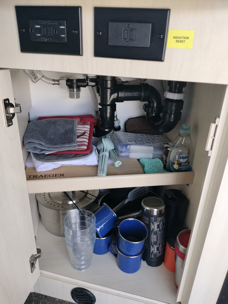 Organized RV rental kitchen cabinet with sink plumbing, electrical outlets, cookware, utensils, and cleaning supplies.