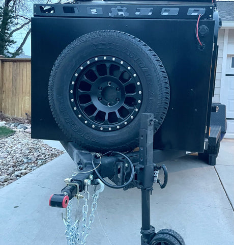 This black, rugged off-road trailer, a versatile RV rental option, features a spare tire, roof rack, and trailer jack. Ready for adventure.