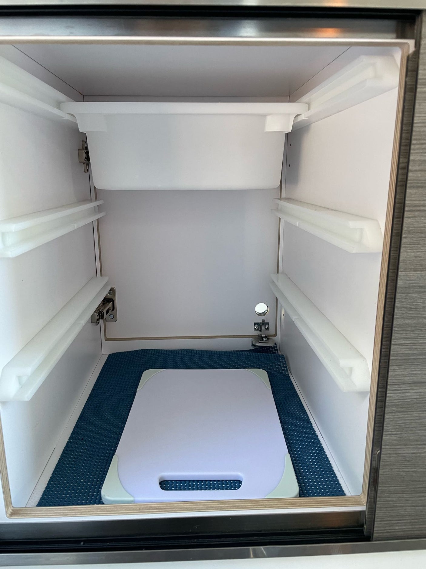 RV rental interior: Clean, empty storage space with white rails, a top bin, and a cutting board on a blue mat.