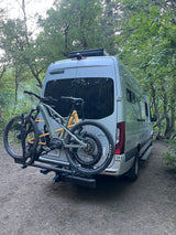 RV rental with bikes ready for forest adventures. A silver camper van and two mountain bikes parked on a dirt path.
