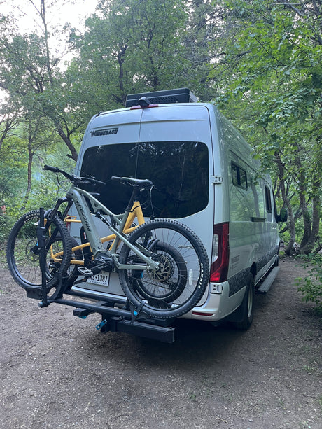 RV rental with bikes ready for forest adventures. A silver camper van and two mountain bikes parked on a dirt path.