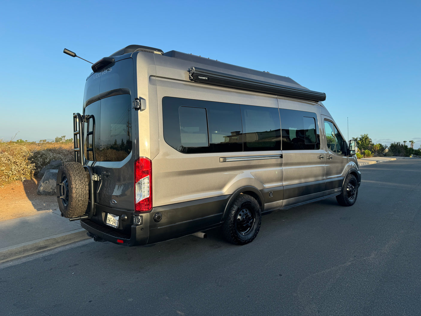 Gray ModVans Ford Transit campervan from side-rear view for rv rental, with off-road tires, ladder & awning.