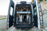 View inside a grey rv rental van from the rear. Open doors show a raised bed, storage units, and a clear aisle to the front.