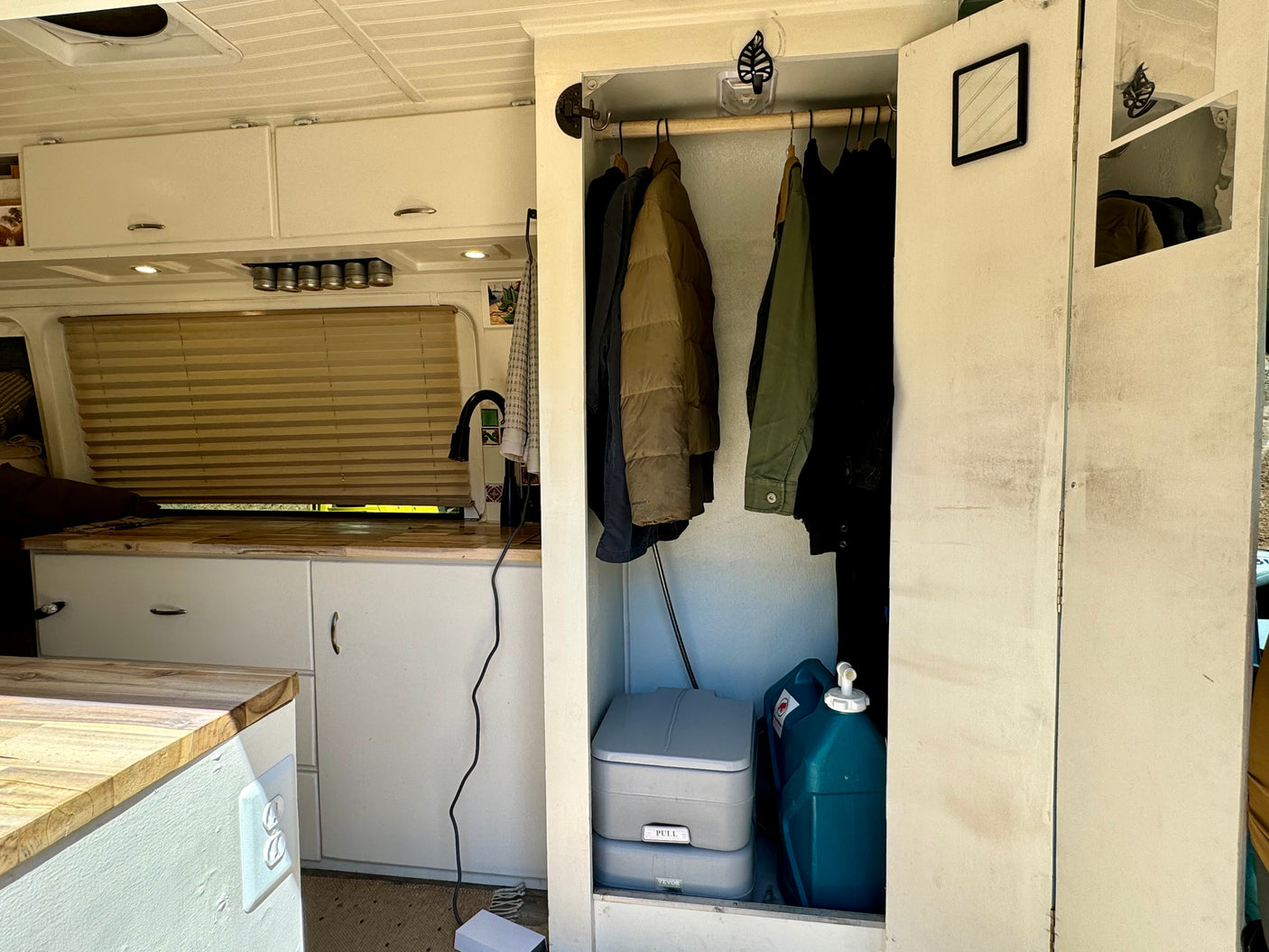 An RV rental's interior: compact kitchenette, wardrobe with hanging clothes, portable toilet, and water jug.