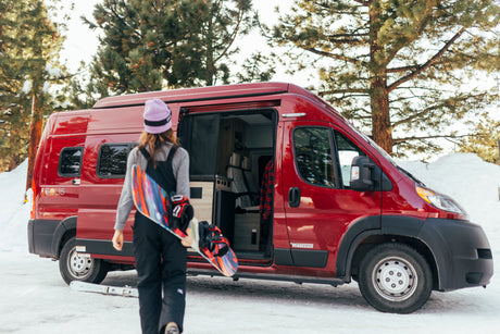 RV rental journey: A snowboarder steps from a red camper van into a snow-covered forest for winter fun.