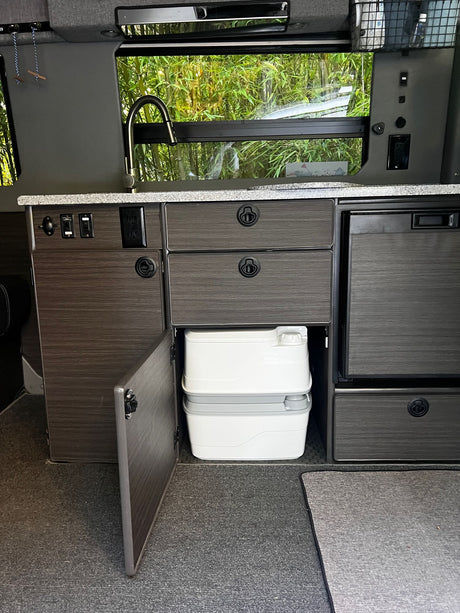RV rental with a compact kitchen setup: sink, faucet, fridge, and a portable toilet stored inside a cabinet. Van life essentials.