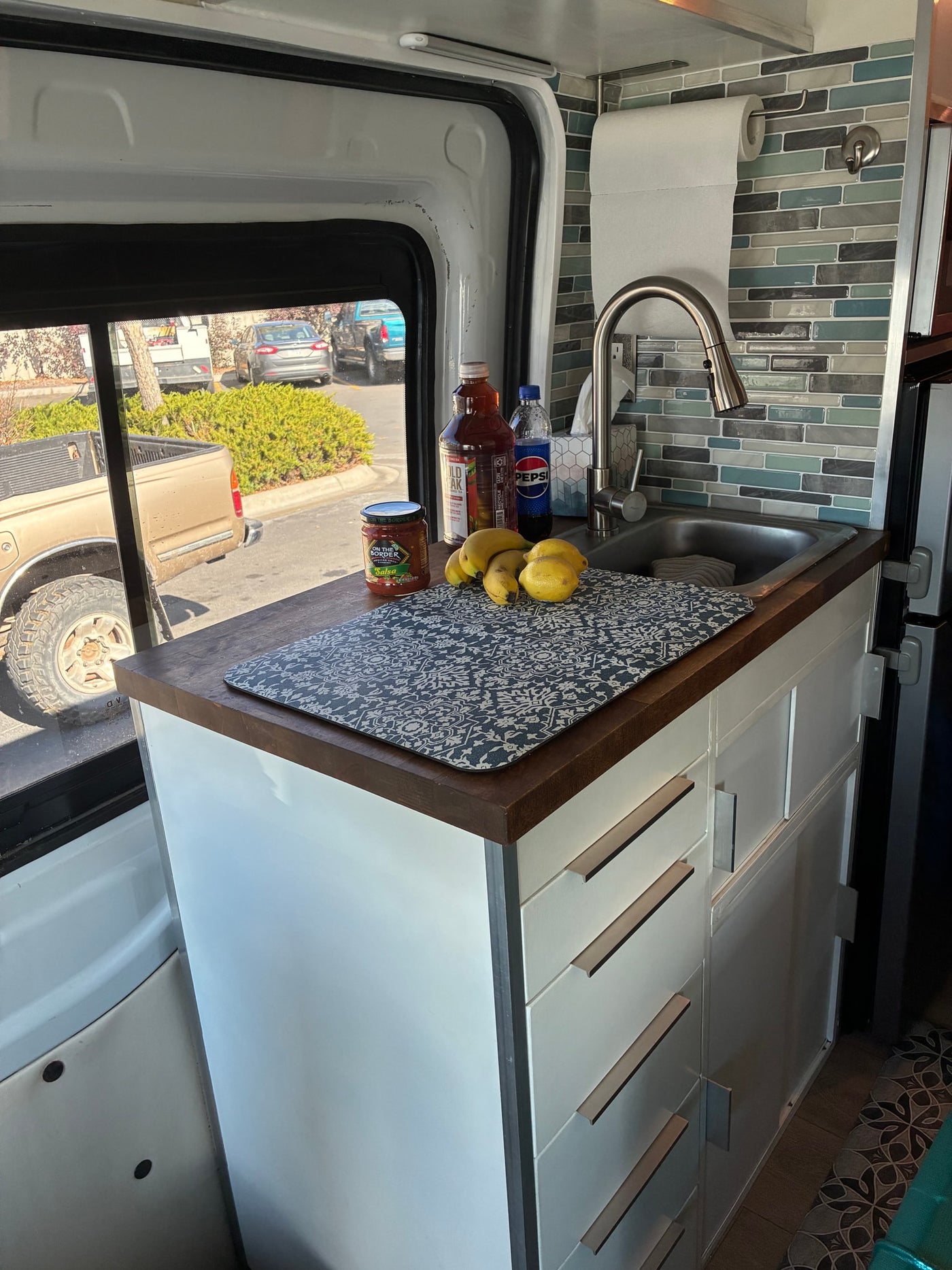 Compact kitchen in an rv rental camper van, with a sink, wooden counter, fruit, and a patterned mat. — photo 5