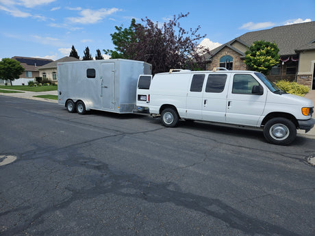 A white van with a silver cargo trailer is parked on a street. This versatile setup, unlike an RV rental, handles large hauls easily.