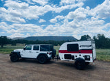Adventure-ready rv rental: White Jeep Willys towing a Sunray Sport camper on a dirt road, mountains & clouds.