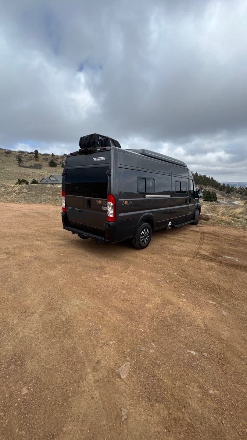 Dark grey camper van (Winnebago Solis) parked on a dirt road under cloudy skies. Perfect for an RV rental adventure.