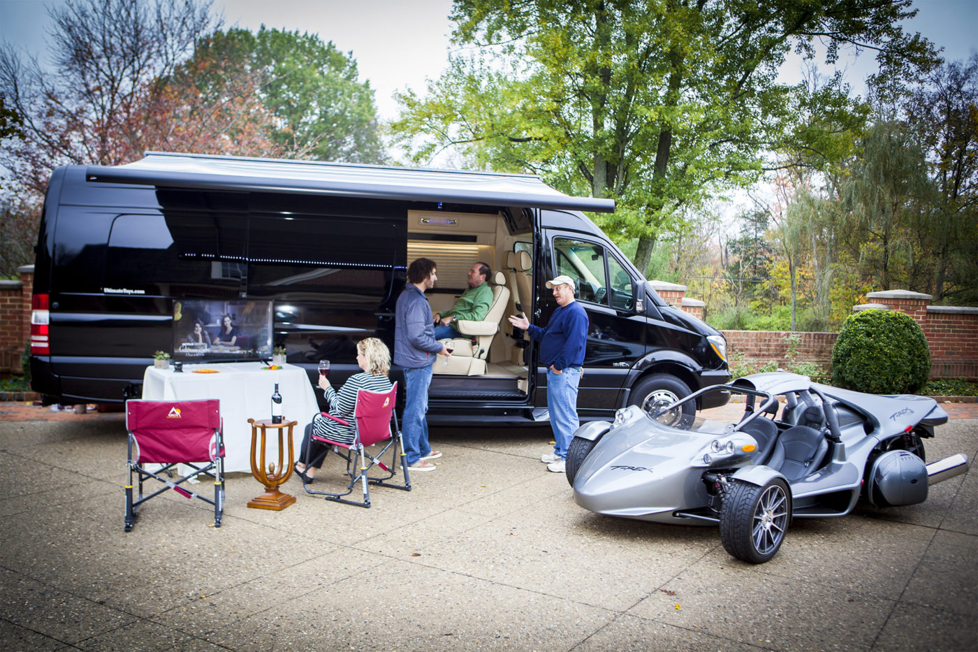 Luxury RV rental scene: Friends gather around a black van with outdoor TV & unique T-Rex vehicle. Social fun!