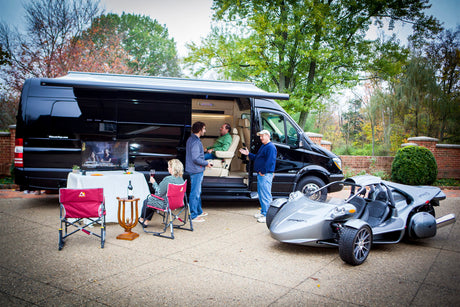 Luxury RV rental scene: Friends gather around a black van with outdoor TV & unique T-Rex vehicle. Social fun!
