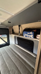 View inside an RV rental: a grey upholstered bed, window showing outdoors, and shelf storage with travel bags.
