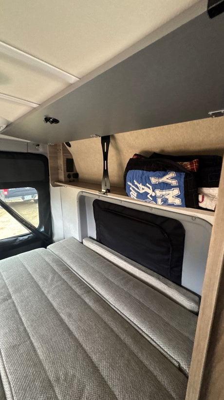 View inside an RV rental: a grey upholstered bed, window showing outdoors, and shelf storage with travel bags.