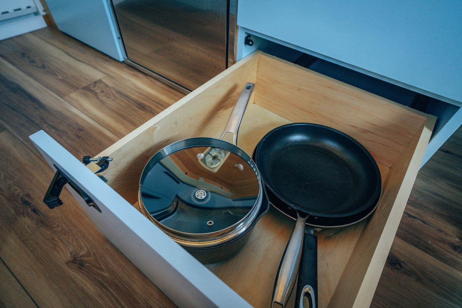 An open kitchen drawer in an RV rental reveals a pot with a lid and a frying pan, on a wooden floor.