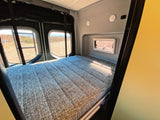 Inside an RV rental: a comfortable bed with a textured gray mattress, flanked by scenic rear windows overlooking a dry landscape.