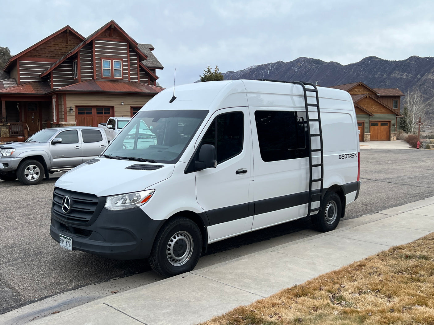 A white Geotrek rv rental van, likely a Mercedes Sprinter, parked on a street with houses and mountains under a cloudy sky.