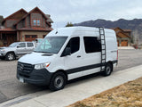 A white Geotrek rv rental van, likely a Mercedes Sprinter, parked on a street with houses and mountains under a cloudy sky.