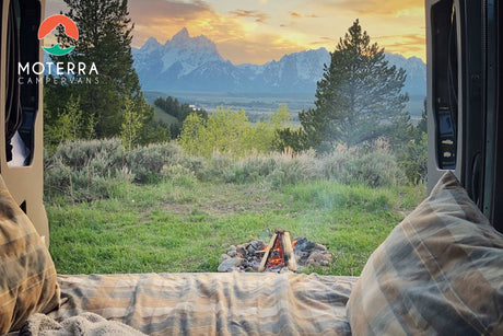 Cozy RV rental view: Inside a camper van, enjoy snow-capped mountains, a sunset sky, and a crackling campfire.