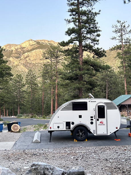 A white Little Guy rv rental teardrop trailer at a scenic mountain campsite, surrounded by towering pine trees.