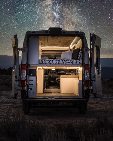 Experience adventure with an RV rental! This camper van, with its lit interior and bed, is parked under a stunning starry night sky.