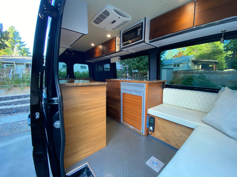  with a bed in back.|RV rental: A person in a green jacket pours water into a coffee maker on a wooden table inside a cozy camper van.|Two people enjoy snacks in their cozy RV rental camper van. Ideal for van life adventures and scenic road trips.|Two people work from a custom rv rental in a desert. Open back doors reveal bed — photo 9