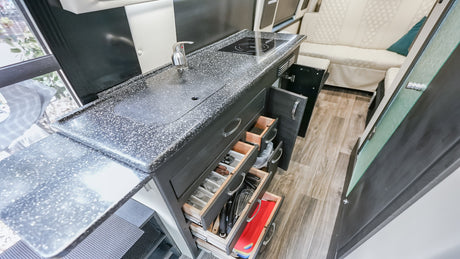 Inside an RV rental, a modern kitchen area with a sink, cooktop, and multiple open drawers showing organized utensils.