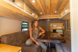 Enjoy this RV rental's inviting interior! A woman relaxes on a bench seat near a dark table in a warm, wood-finished space.