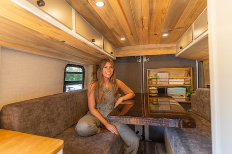 Enjoy this RV rental's inviting interior! A woman relaxes on a bench seat near a dark table in a warm, wood-finished space.