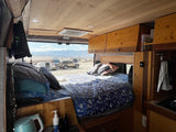 Cozy RV rental interior with a bed and wooden accents, overlooking a desert campsite and mountains.