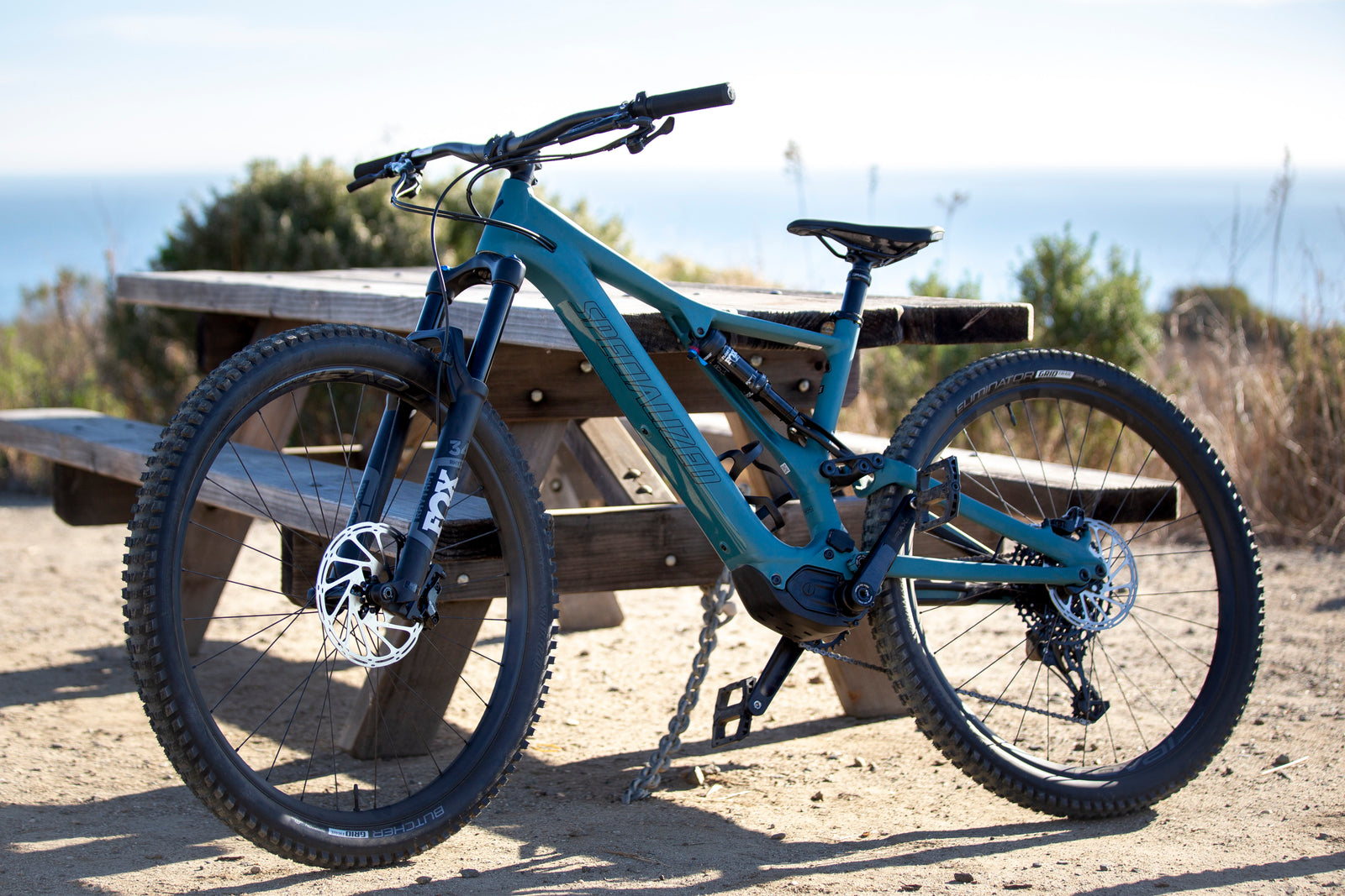 A teal Specialized e-mountain bike is parked by a picnic table, with an ocean view in the background. Perfect for RV rental adventures.