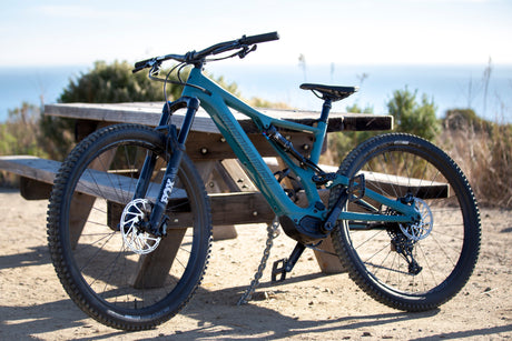 A teal Specialized e-mountain bike is parked by a picnic table, with an ocean view in the background. Perfect for RV rental adventures.
