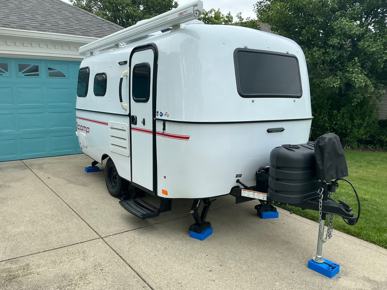 A compact white RV rental with a Fiamma awning on top, parked on a driveway with blue leveling blocks.