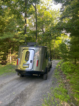 A shiny Airstream rv rental navigates a gravel forest road, surrounded by lush green trees and ferns on a sunny day.
