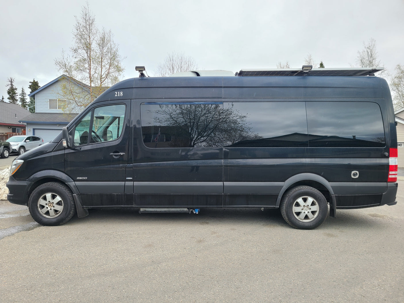 Black Mercedes-Benz Sprinter RV rental, marked 218, with solar panels, parked in a suburban setting.
