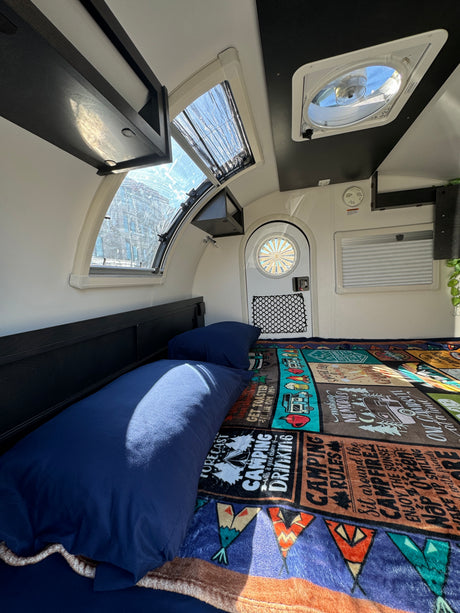Inside a compact rv rental camper with a bed, "Camping Rules" blanket, bright windows, overhead storage, and a ceiling fan.