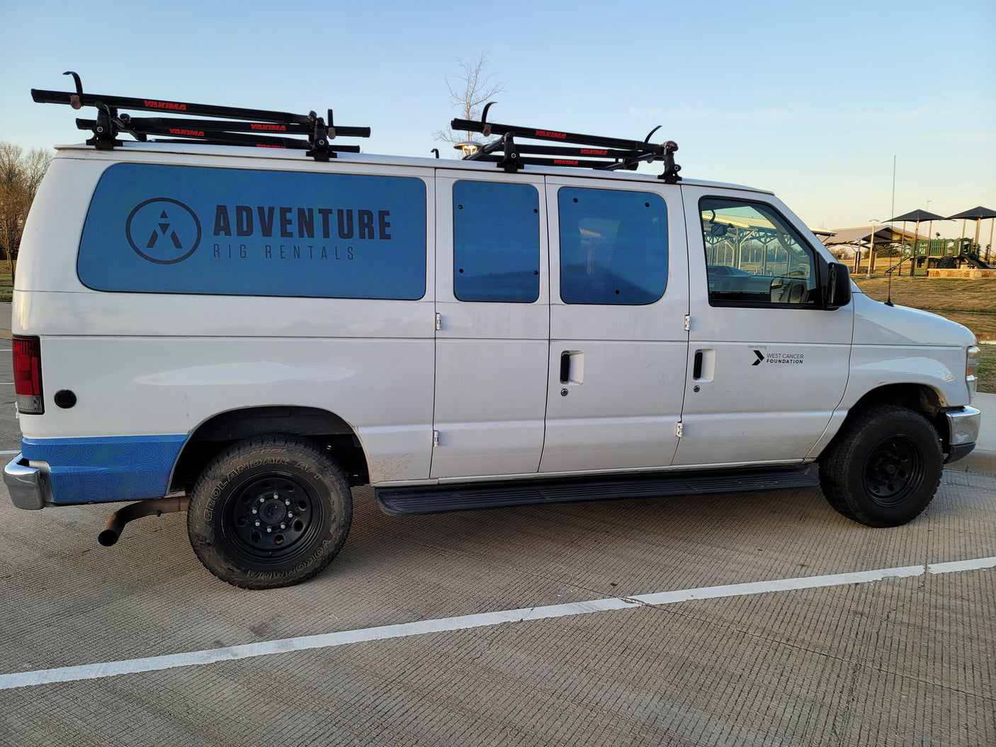 White "Adventure Rig Rentals" van, an RV rental, with Yakima roof racks, benefiting West Cancer Foundation.
