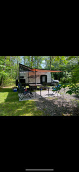 An RV rental, a Viking travel trailer, set up in a wooded campsite with an orange awning, chairs, and tables.