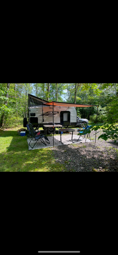 An RV rental, a Viking travel trailer, set up in a wooded campsite with an orange awning, chairs, and tables.