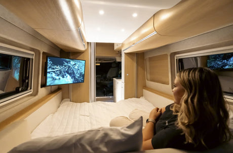 Inside a modern RV rental, a woman with long hair lies on the comfy bed watching TV. Wooden walls & ceiling add warmth.