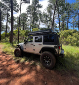 White Jeep Wrangler with rooftop tent on a dirt path in a pine forest, ideal for rv rental adventures and off-road trips.