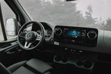 Interior of a Mercedes rv rental: dashboard, steering wheel, and infotainment system. Rain spatters the windshield, showing blurred trees.