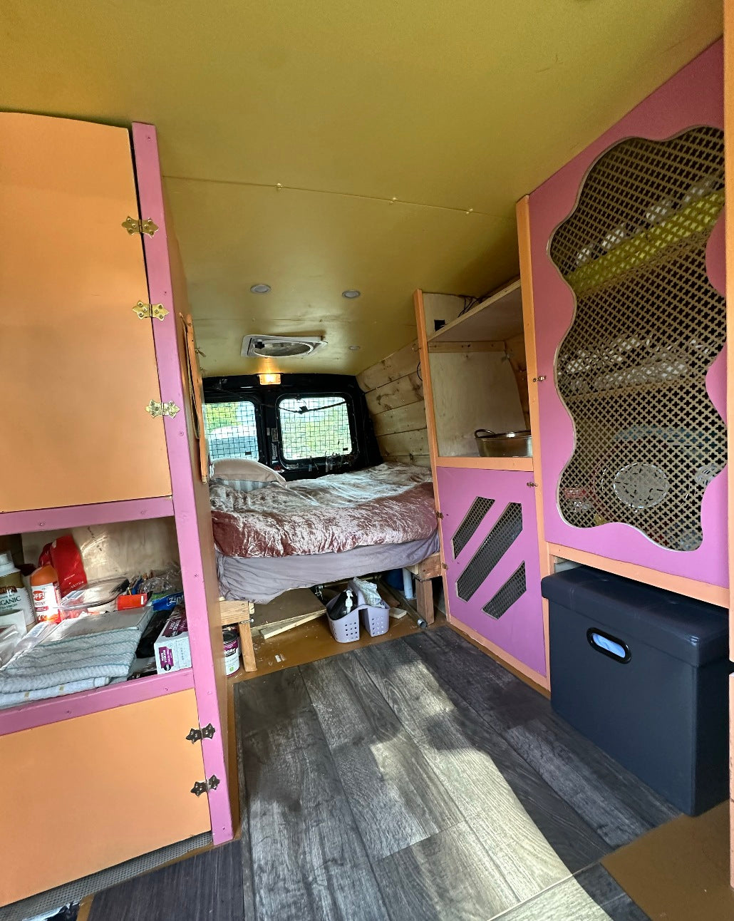 Cozy RV rental camper van interior. Features a bed, vibrant pink & orange custom cabinetry, and wood-look flooring.
