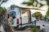 This rv rental shows a light camper van with an open side, extended awning, and outdoor cooking area at a lively campsite.